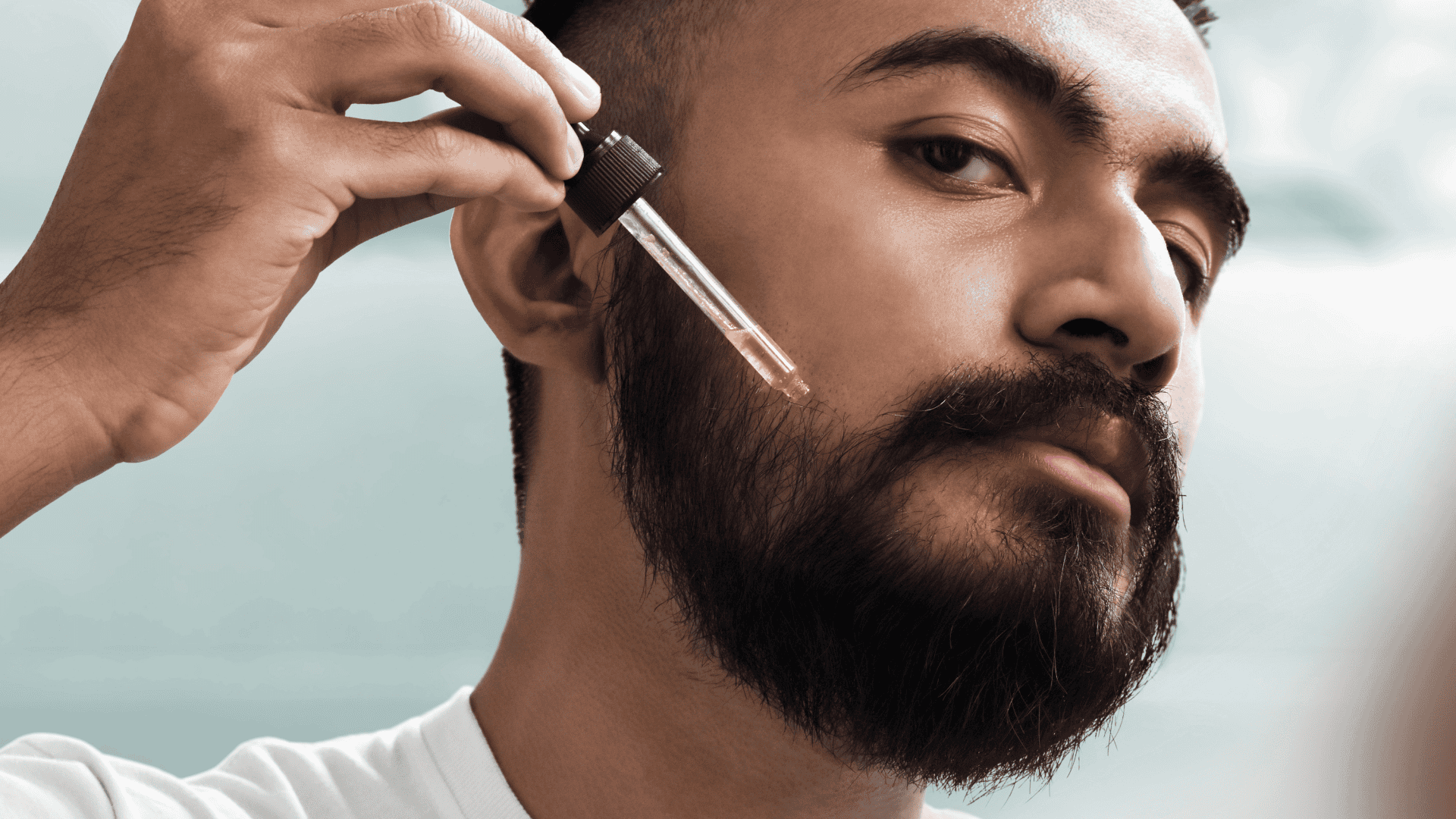 Man applying a drop of beard oil to his beard against a light blue background